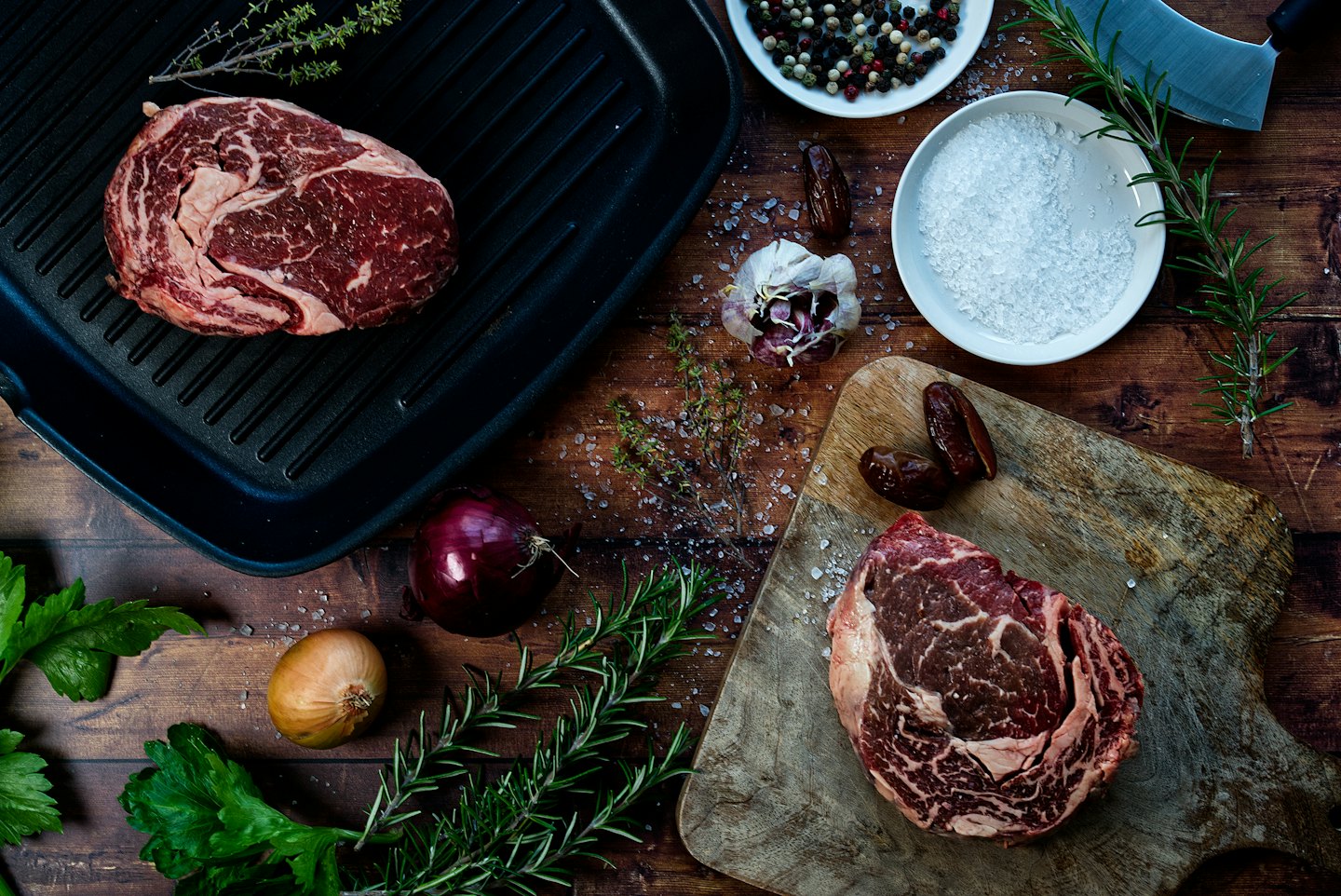 Two raw ribeye steaks with herbs and seasonings for cooking.